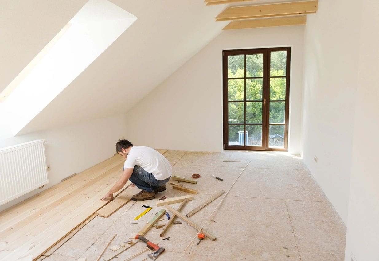 A man is laying down wood in an unfinished room.